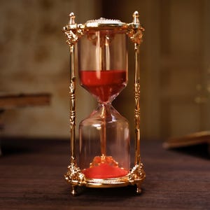 Può includere: Una clessidra dorata con un timer di sabbia rosso. La clessidra è su una superficie di legno.