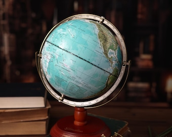 Op de afbeelding: Een tafelglobe met lichtblauwe oceanen en groene landmassa's, gemonteerd op een messingkleurige meridiaanring met graadmarkeringen. De globe rust op een donkerbruine houten voet met het woord Timeless in de voorzijde gegraveerd.