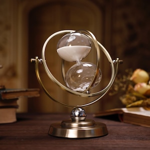 Può includere: Una clessidra dorata con una clessidra di sabbia bianca all'interno è montata su un supporto decorativo. Il supporto ha una base rotonda e una cornice circolare che tiene la clessidra. La clessidra è su un tavolo di legno con libri e fiori sullo sfondo.