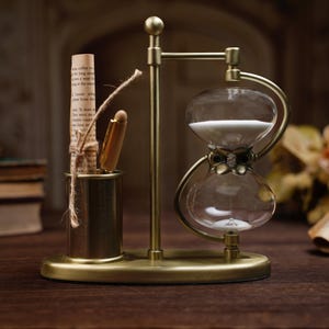 May include: An antique brass hourglass with a pen and a rolled-up paper scroll. The hourglass has a clear glass bulb filled with white sand. The pen and scroll are in a small brass holder. The scene is set on a wooden surface.