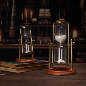 Könnte beinhalten: Zwei antike Sanduhren aus Messing auf Holzsockeln. Die größere Sanduhr hat weißen Sand und die kleinere Sanduhr hat klaren Sand.