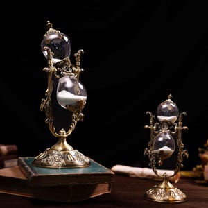 May include: Two ornate gold-toned hourglass sand timers with clear glass bulbs. The larger hourglass is on a book, while the smaller hourglass is on a wooden surface.