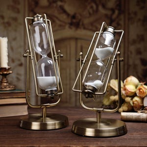 Könnte beinhalten: Zwei antike Sanduhren mit transparenten Glaskammern und weißem Sand. Die Sanduhren sind in einem messingfarbenen Metallrahmen und -sockel angeordnet. Das Design ist vintage, mit einer klassischen Ästhetik. Der Hintergrund umfasst eine Kerze und Bücher.