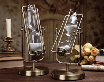 Op de afbeelding: Twee antieke zandlopers met heldere glazen kamers en wit zand. De zandlopers staan in een messingkleurig metalen frame en voet. Het ontwerp is vintage, met een klassieke esthetiek. De achtergrond bevat een kaars en boeken.