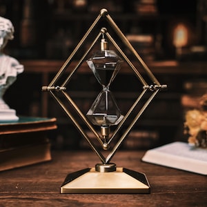 Könnte beinhalten: Dekorative Sanduhr mit goldfarbenem, rautenförmigem Rahmen. Die Glasbehälter enthalten dunklen Sand. Die Sanduhr steht auf einer Holzoberfläche. Der Hintergrund umfasst eine Büste, Bücher und Trockenblumen. Maße in Zentimetern.