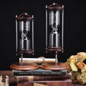 May include: Two wooden hourglasses with clear glass chambers and silver metal supports. Each hourglass has a dark wood base and top, with engraved text. One reads "Mr & Mrs Jackson" and the other "Siren & Ankit".