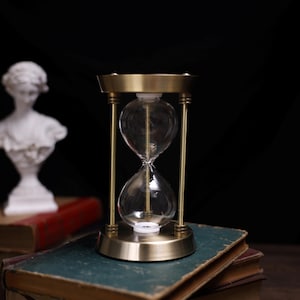 May include: A brass hourglass with a glass bulb sits on a stack of old books. The hourglass is on a wooden surface.