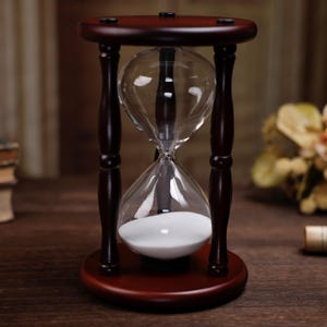 Könnte beinhalten: Eine klassische Sanduhr mit einem dunkelbraunen Holzrahmen und -sockel. Die klare Glaskugel enthält weißen Sand, der die Zeit symbolisiert. Diese dekorative Sanduhr ist ein stilvolles Wohnaccessoire oder ein durchdachtes Geschenk.