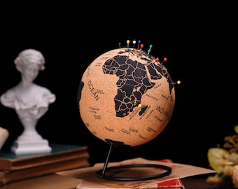 May include: A cork world globe with a black map and colorful push pins. The globe is on a black metal stand and is sitting on a stack of books.