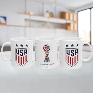 May include: White ceramic mug featuring the USA soccer team logo with red and blue stripes and three stars. Another mug displays the 2026 FIFA World Cup logo with the text "Mexico USA Canada".