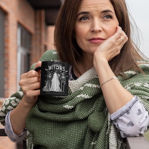 May include: Black ceramic mug with the text "Drink up Witches" and a graphic of three figures in white. The mug is held by a person wearing a green and white patterned shawl. The mug is a Halloween-themed product.