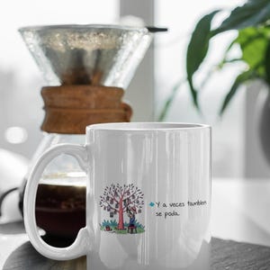 May include: White ceramic mug with a whimsical illustration of a tree with hanging lanterns and a person. The mug has a large handle and the text "Y a veces tambien se poda."