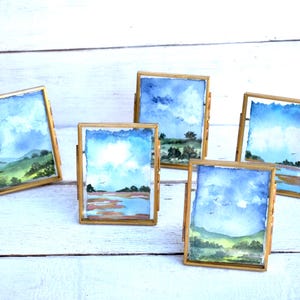 May include: Five rectangular brass frames containing small watercolor landscape paintings. Each frame features a unique scene with blue skies, white clouds, green hills, and water elements, all displayed in a vertical orientation with clear glass.