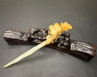 May include: A decorative hair stick with a floral design. The stick is pale yellow and features a cluster of orange flowers at the top. It rests on a dark brown, carved wooden base.