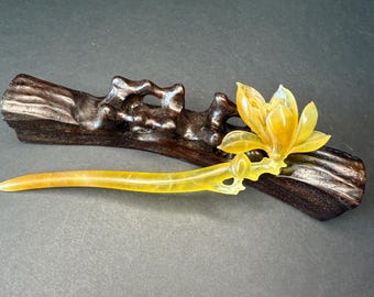 May include: A yellow lotus flower hair stick with a carved floral design. The hair stick is displayed on a dark brown wooden stand with a natural, organic shape. The hair accessory is a decorative item.