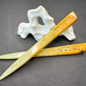 Puede incluir: Dos abridores de cartas con diseños florales tallados, en tonos amarillos y marrones, se exhiben sobre una superficie oscura. Una escultura de cerámica blanca y abstracta está en el fondo.