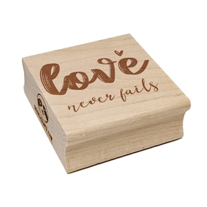 May include: A wooden stamp with the words "love never fails" in brown script. The stamp has a rectangular shape with a beveled edge and a small logo on the side. The stamp is made of light-colored wood.