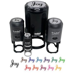 May include: A collection of black self-inking stamps with the word "Joy" in a script font. The stamps are various sizes, with the brand name "Trodat" visible. Below the stamps, the word "Joy" is repeated in multiple colors.