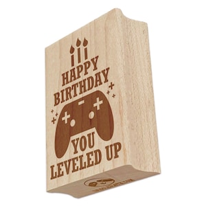 May include: A wooden stamp with the text "HAPPY BIRTHDAY YOU LEVELED UP" and a game controller graphic. The stamp has a light brown color and features three candle illustrations at the top. The design is in a darker brown color.