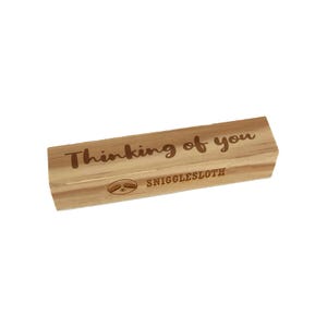 May include: A rectangular wooden block with the text "Thinking of you" in a handwritten style. Below the text is the word "SNIGGLESLOTH" and a sloth illustration. The wood has a light brown color.