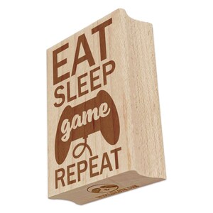 May include: A wooden stamp with the words "EAT SLEEP game REPEAT" and a game controller graphic. The text and graphic are in a dark brown color, and the stamp is made of light-colored wood.