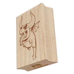 May include: A wooden stamp featuring a cartoon devil with horns, wings, and a long tongue. The stamp has a light brown color and a curved handle. The devil is drawn in a simple, cartoon style.