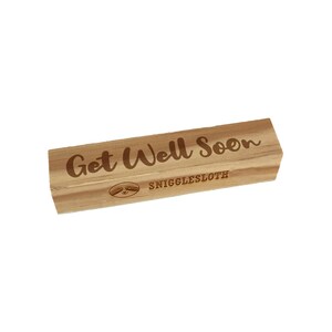 May include: A rectangular wooden stamp with the words "Get Well Soon" engraved in a cursive font. Below the text is the word "SNIGGLESLOTH" and a sloth illustration. The stamp is light brown.