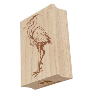 May include: A wooden stamp with a detailed line drawing of a heron standing on one leg. The stamp is made by "The Sloth Hut" and is a light brown color.