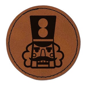 May include: A circular brown leather patch featuring a black laser-engraved graphic of a nutcracker. The design shows a stylized nutcracker with a tall hat, a prominent mustache, and a square jaw, finished with black stitching around the edge.