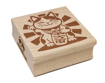 Op de afbeelding: Een vierkante houten rubberstempel met een lasergegraveerd ontwerp van een wenkende kat, bekend als maneki-neko, die een munt met Japanse karakters vasthoudt. De kat is gecentreerd tegen een zonnestraalpatroon en de zijkant toont een klein luiaardlogo.