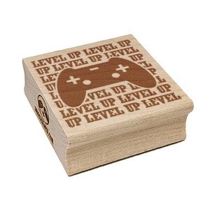 May include: A wooden stamp with the text "LEVEL UP" repeated around a brown controller graphic. The stamp is rectangular and has a light wood finish. The controller graphic has a plus sign and two buttons.