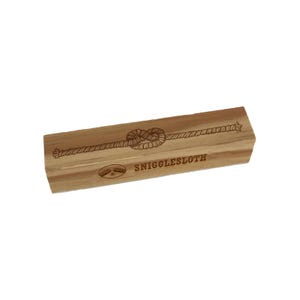 May include: A rectangular wooden stamp with a rope knot design and the text "SNIGGLESLOTH". The stamp is light brown with a detailed engraving of a rope knot in the center and a sloth icon.