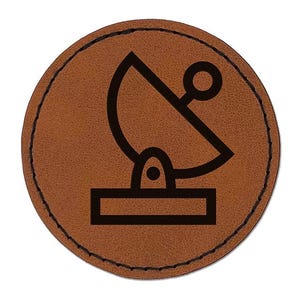 May include: A circular brown patch with a dark brown stitched border. The center features a black graphic icon of a satellite dish mounted on a rectangular base, depicted with a simple line drawing style on a textured, leather-like surface.