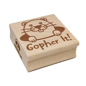 May include: A wooden stamp with a cartoon gopher design and the phrase "Gopher It!" in brown ink. The stamp has a rectangular shape with rounded edges and a light wood color. The gopher is smiling and peeking out of a hole.