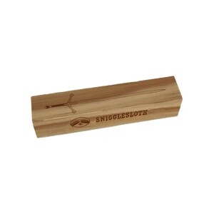 May include: A rectangular wooden box with a sword design and the text "SNIGGLESLOTH" engraved on the surface. The sword is depicted with a cross-shaped hilt. The box is light brown.