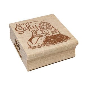 May include: A wooden stamp with the words "Always Salty" and an illustration of a mermaid. The mermaid is sitting on a rock, with her tail in the water. The stamp is made of light-colored wood.