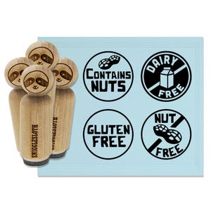 May include: Wooden stamps with sloth faces and the text "SNIGGLESLOTH" are next to a light blue card with black and white circular food allergy labels that read "CONTAINS NUTS", "DAIRY FREE", "GLUTEN FREE", and "NUT FREE".