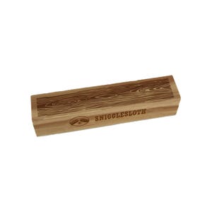 May include: A rectangular wooden block with a wood grain pattern on the top surface. The block has the word "SNIGGLESLOTH" printed on the side, along with a small graphic of two sloths. The block is light brown.