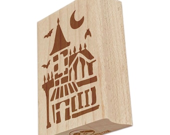 May include: A rectangular wooden rubber stamp featuring a laser-engraved design of a tall house with a pointed roof, a crescent moon, and two flying bats. The light-colored wood block has a smooth finish and a small logo printed on the bottom edge.