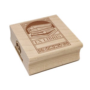 May include: A wooden stamp with the words "EX LIBRIS" and an illustration of stacked books. The stamp has a rectangular design with decorative borders and is made of light-colored wood.