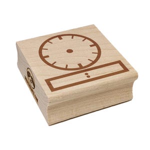 May include: A wooden stamp with a clock face design and a rectangular box below it. The clock face has a circular shape with brown markings for the hours. The rectangular box has two dots inside. The stamp is made of light-colored wood.