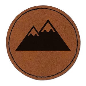 May include: A circular brown leather patch featuring a black graphic of two mountain peaks. The patch has a smooth texture and is finished with a dark stitched border that runs along the entire circumference of the circular leather piece.