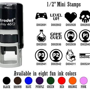 May include: A black and white self-inking stamp with the text "trodat printy 4612" and a variety of 1/2 inch mini stamps with game-themed designs. The designs include a joystick, a heart, a skull, a potion bottle, the words "Game Over", a player icon with the number "4", a player icon with the number "1", a player icon with the number "3", a player icon with the number "2", a bunny rabbit with headphones, and the word "Loading..." The stamps are available in eight colors: black, brown, blue, purple, green, red, orange, and pink.