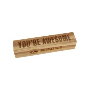 May include: A rectangular wooden block with the words "YOU'RE AWESOME" engraved in a bold, sans-serif font. Below the text is the word "SNIGGLESLOTH" and a sloth graphic. The wood has a light brown color.