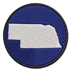 May include: A circular embroidered patch featuring a white silhouette of the state of Nebraska centered on a solid royal blue background. The patch is finished with a thick black stitched border around the entire outer edge of the circle.