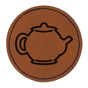 May include: A circular brown leather patch featuring a black outline of a teapot in the center. The patch has a stitched border around the edge, creating a defined circular shape with a smooth, textured surface and a clear, dark graphic design.