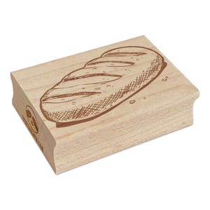 May include: A wooden stamp featuring a detailed illustration of a loaf of bread. The bread is depicted with cross-hatched lines and brown ink, with visible texture and scoring. The stamp has a rectangular shape with rounded edges and a logo on the side.
