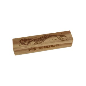May include: A rectangular wooden block with a mermaid illustration and the text "SNIGGLESLOTH" engraved on it. The mermaid has long hair and a patterned tail. Small fish are also engraved on the block.