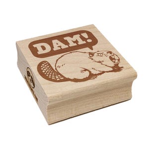 May include: A wooden stamp featuring a beaver with the word "DAM!" in a speech bubble. The beaver is brown, with an open mouth and a large tail. The stamp is made of light-colored wood.