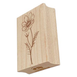 May include: A wooden stamp featuring a detailed illustration of a cosmos flower. The flower and stem are depicted in a brown color, set against the natural wood grain. The stamp has a curved handle for easy use.